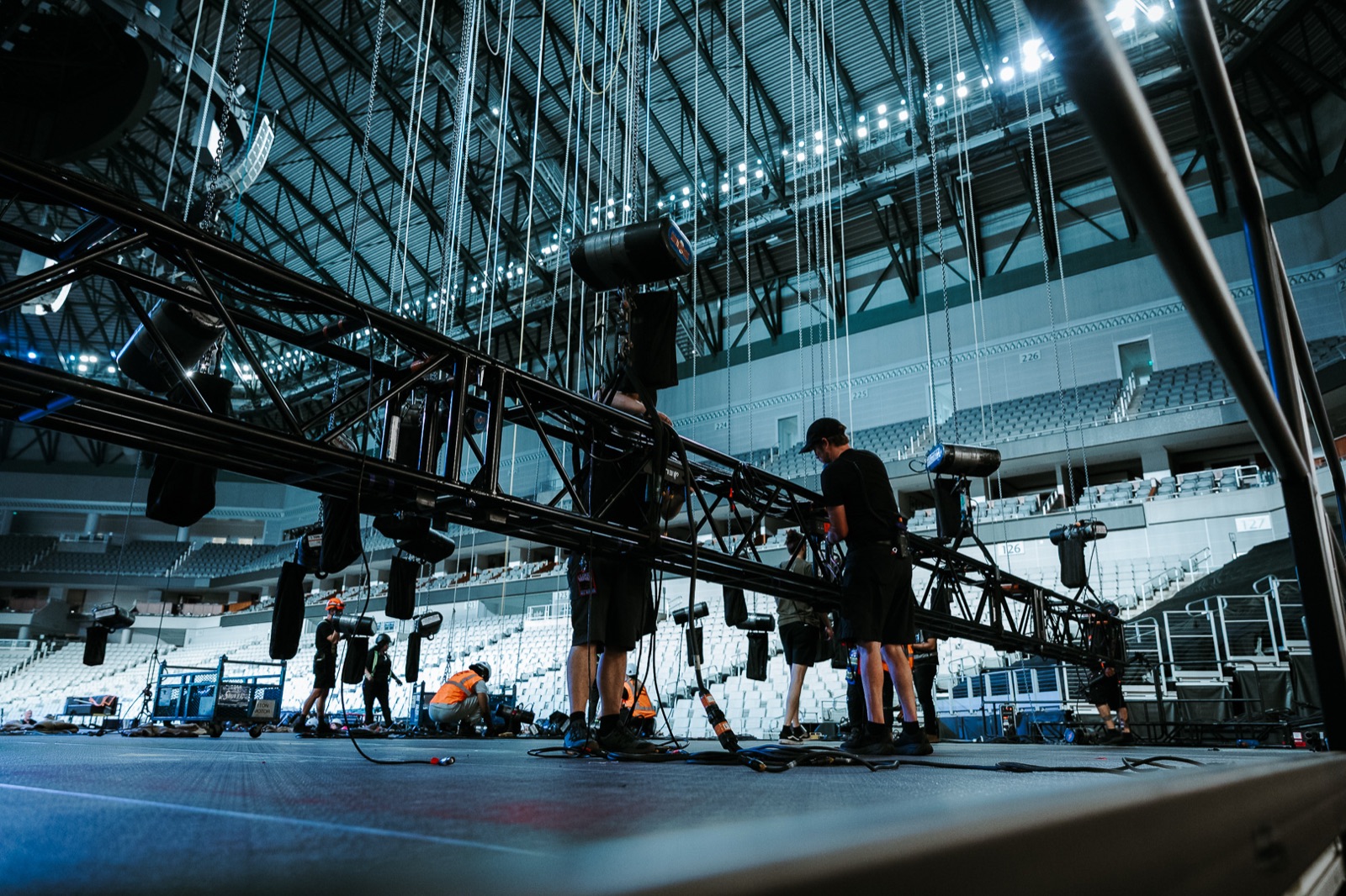 Crew building lighting truss on the arena floor, empty seating bowl behind them